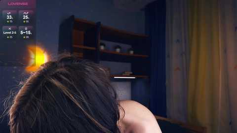 Media: Video of a woman with long, dark hair, seen from behind, standing in a dimly lit, dark blue room with shelves and a bed, displaying a virtual reality game overlay.