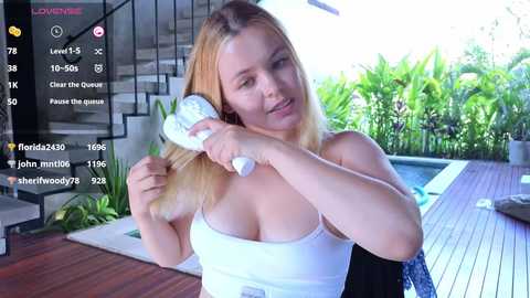 Media: Video of a blonde woman with fair skin, medium build, medium breasts, wearing a white tank top, holding a hairdryer, in a modern, sunlit room with a pool.