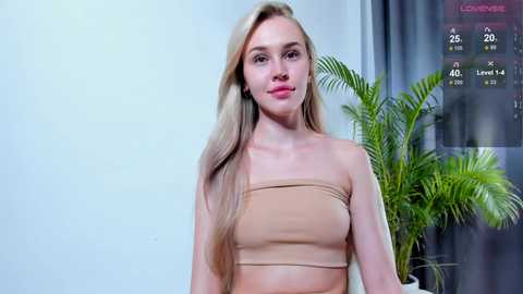 Media: Video of a slender, light-skinned woman with long, straight blonde hair, wearing a beige strapless top. She stands in a modern indoor setting with a large potted palm plant behind her.