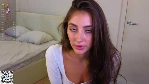 Media: Video of a young woman with long brown hair, wearing a white top, sitting on a bed in a simple, modern bedroom.