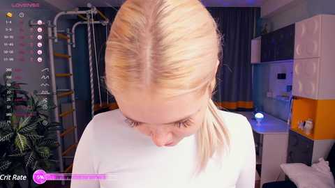 Media: Video of a blonde woman with straight hair, wearing a white top, in a modern, colorful bedroom with gym equipment and potted plants.