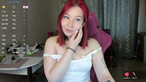 Media: Video of a young woman with red hair, wearing a white off-shoulder top, sitting in a pink gaming chair. Background includes a desk cluttered with papers, a window with string lights, and a room with a carpeted floor.