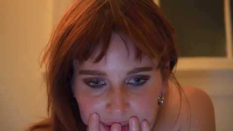 Media: Video of a young woman with fair skin, long auburn hair, and dark eyeliner, looking downward, holding her fingers to her lips, in a softly lit indoor setting.