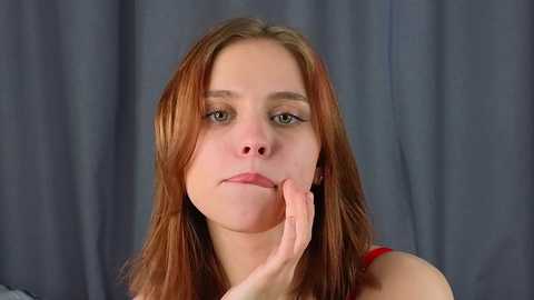 Media: Video of a young Caucasian woman with fair skin, straight red hair, and green eyes. She has a neutral expression, with one hand gently touching her chin. She wears a red top. Background is a plain grey curtain.