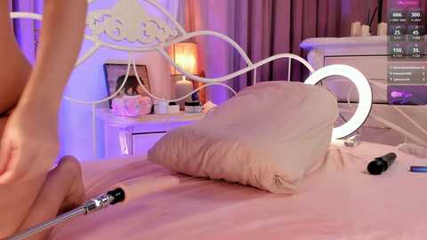 Media: Video of a woman's legs on a bed with white metal headboard, pink sheets, and a large pillow. She's adjusting a sex toy, with a TV screen showing a live stream of the scene.