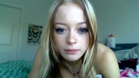 Media: A video of a young, fair-skinned blonde woman with blue eyes and a nose ring, wearing a black top, standing in a cluttered bedroom with a white door, a bed, and a \"PAB\" can.