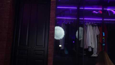 Media: Video of a dimly-lit room with a brick wall, black doors, and a glass wardrobe illuminated by purple LED lights, displaying hanging clothes and shoes.