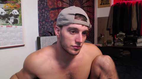 Media: Video of a shirtless, muscular, fair-skinned man with short brown hair, wearing a backward gray baseball cap, indoors with a colorful tapestry and calendar behind him.