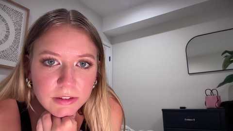 Media: Video of a Caucasian woman with light skin and straight blonde hair, wearing hoop earrings and a black top, looking contemplative. Background includes a white wall, a black dresser with a pink bag, and a round mirror.