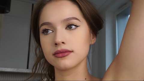 Media: Video of a young woman with light skin, brown hair, and blue eyes, looking introspective in a modern kitchen. She has light makeup, including mascara, and wears a yellow top.