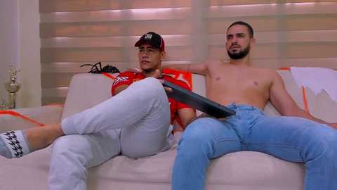 Media: Video of two shirtless men, one in a red baseball cap, white pants, and black shoes, the other in blue jeans, playing with a remote control on a beige sofa.