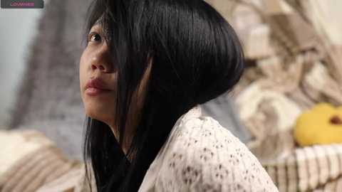 Media: Video of an East Asian woman with a sleek, black, asymmetrical bob haircut, wearing a light-colored knitted sweater. Background features a beige, patterned couch with a yellow fruit.