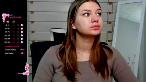 Media: A video of a young woman with straight brown hair, wearing a brown sweater, gazing thoughtfully. Background features a white wall and window blinds.