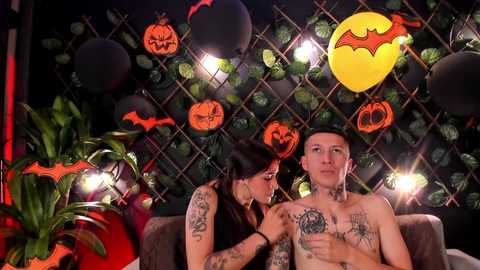 Media: Video of a tattooed woman kissing a shirtless man with a Batman balloon in a Halloween-themed room with jack-o'-lanterns and spider decorations.