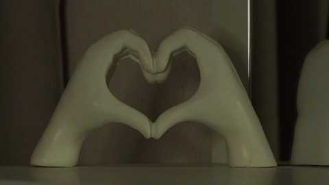 Media: A video of two white ceramic hands forming a heart shape on a shelf, against a dark, muted background. The hands are smooth and slightly glossy, creating a soft, warm light reflection.