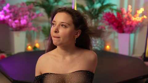 Media: Video of a fair-skinned woman with long brown hair, wearing a black sequined off-shoulder dress, standing against a backdrop of vibrant pink flowers and lit candles.