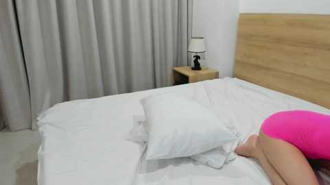 Media: Video of a woman in a bright pink swimsuit lying on a white bed with white pillows, against a white wall and wooden headboard, with a wooden nightstand and lamp.