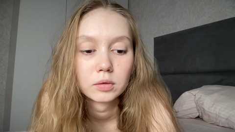 Media: Video of a young Caucasian woman with long, wavy blonde hair and pale skin, looking sad and downcast, sitting on a bed with white pillows in a gray-toned, minimalist bedroom.