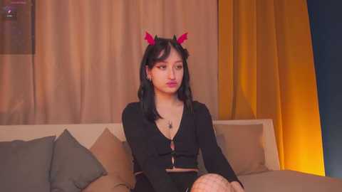 Media: Video of an Asian woman with long black hair and red devil horns, wearing a black top, sitting on a beige sofa, with a neutral-toned background.