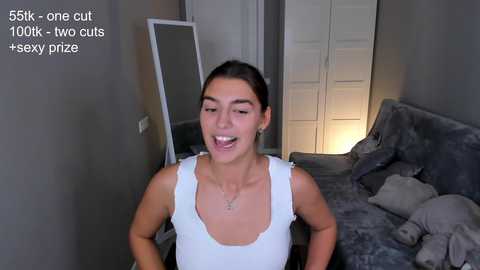 Media: A video of a young woman with medium skin tone and dark hair, wearing a white tank top, smiling, standing in a modern, gray-toned bedroom.