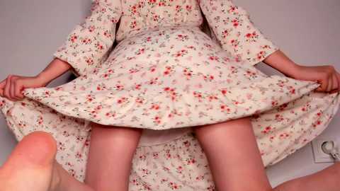 Media: Video of a woman in a floral dress, lifted by a man's hands, revealing her bare thighs and feet. The background is a plain white wall with a light switch.