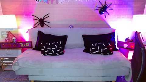 Media: Video of a cozy, Halloween-themed bedroom with a white sofa adorned with black pillows featuring white ghost designs. Spider decorations hang from the wall, and colorful fairy lights and a nightstand with a lamp are visible.