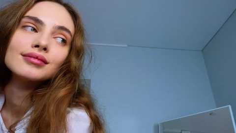 Media: A video of a young woman with long, wavy brown hair, fair skin, and light makeup, looking upward thoughtfully. She wears a white top. Background: white walls, ceiling, and a mirror.