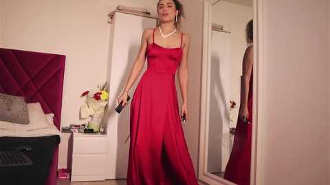 Media: Video of a confident woman in a red satin gown, standing in a modern bedroom with white walls, a bed, and a mirror.