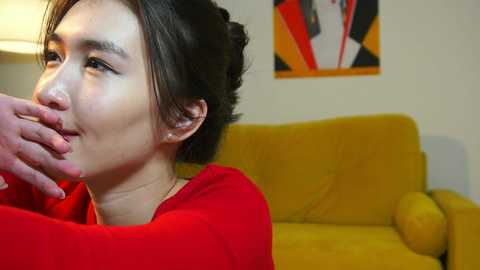 Media: Video of a young East Asian woman with short black hair, wearing a red sweater, sitting on a mustard-yellow sofa in a cozy living room.