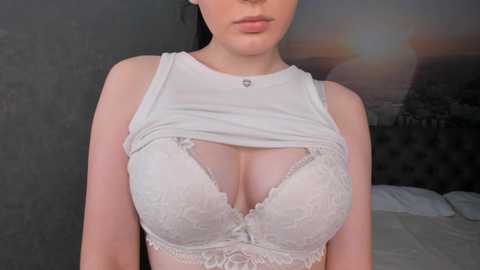 Media: Video of a fair-skinned woman with dark hair, wearing a white lace bra that accentuates her large breasts, against a dark, textured wall background.