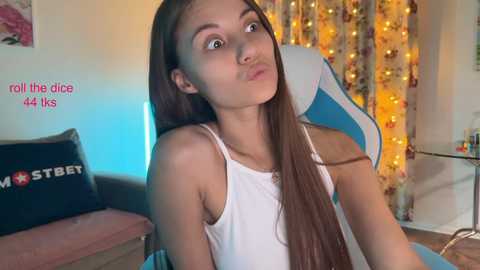 Media: A video of a young woman with long brown hair, wearing a white tank top, sitting in a blue gaming chair. She has a neutral expression and a slight pout. The background features a colorful, cozy room with fairy lights and a floral pillow.