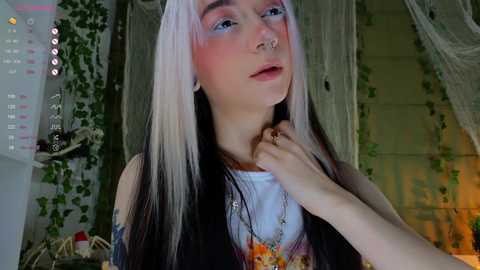 Media: Video of a young woman with long, pastel-colored hair, wearing a white T-shirt, sitting in a cozy room with greenery and drapes.