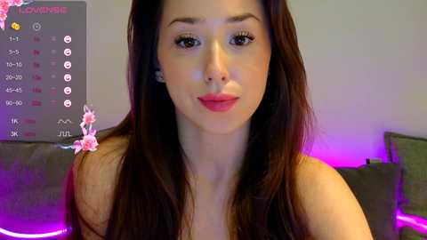 Media: Video of a light-skinned woman with long, straight brown hair, wearing red lipstick, sitting on a gray couch, in a room with purple and pink ambient lighting, with a calendar and pink flower overlay.