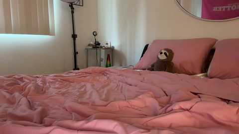 Media: A video of a cozy bedroom with a pink bedspread, a stuffed sloth on a pillow, a nightstand with a lamp and books, and a round mirror on the wall.