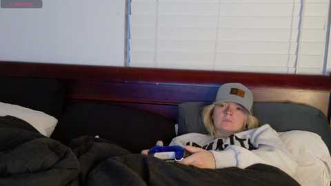 Media: Video of a blonde woman in a gray baseball cap, gray hoodie, and black blanket, lying on a dark wooden bed in a dimly lit room with white blinds.