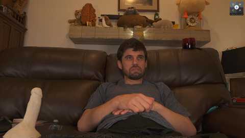 Media: Video of a man with short brown hair and beard, wearing a gray shirt, sitting on a brown leather couch, holding a white stuffed animal, in a cozy living room with a fireplace mantle, framed art, and decorative items.