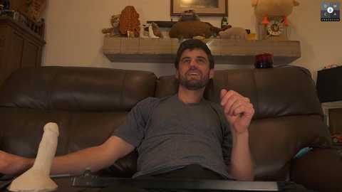 Media: Video of a bearded man in a gray T-shirt, reclining on a brown leather couch, holding a white plastic toy, in a warmly lit living room with a TV, wooden furniture, and decorative items.