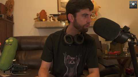 Media: Video of a bearded man with headphones, wearing a black graphic T-shirt, sitting in a cozy living room with a brown couch, microphone, and toy figurines.