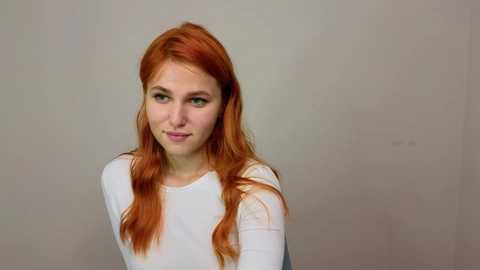 Media: Video of a fair-skinned young woman with long, wavy, bright red hair, wearing a white long-sleeved shirt, against a plain, light-colored wall background. She looks slightly to the side with a neutral expression.