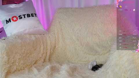 Media: Video of a plush, cream-colored bed with a white pillow featuring \"MOSTBET\" in bold red letters. Soft, fuzzy blankets cover the bed, with a black and white toy in the lower right corner.