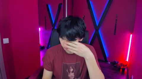 Media: Video of a young man with short, dark hair, wearing a maroon t-shirt with a graphic, sitting in a chair, covering his face with his right hand, in a dimly lit room with red and blue neon lights, BDSM-themed decor.