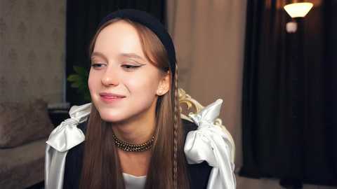 Media: Video of a young Caucasian woman with light skin, long brown hair, wearing a black beret, white blouse with puffed sleeves, and a gold chain necklace, smiling slightly, in a dimly-lit room with beige walls and dark curtains.