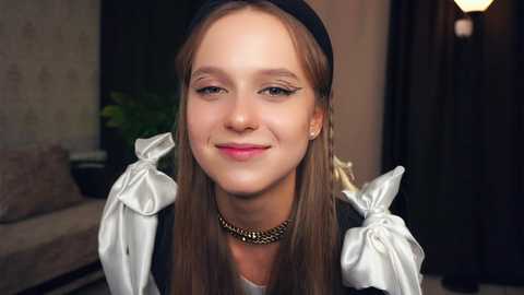 Media: Video of a young, light-skinned girl with long brown hair, wearing a maid outfit with white ruffled sleeves, a black headband, and a gold chain necklace, smiling softly. Background includes a sofa and green plant.