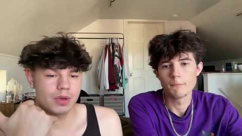 Media: Video of two young men with curly hair in a bedroom. One wears a black tank top, the other a purple t-shirt with a chain necklace. Closet with hanging clothes in background.