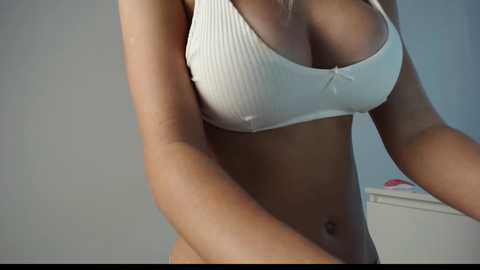 Media: Video of a woman with fair skin and blonde hair, wearing a white ribbed bra with a small bow, highlighting her medium-sized breasts and toned stomach. Background shows a white dresser with a pink object on it.