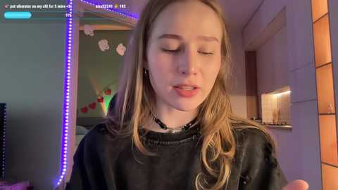 Media: Video of a young woman with long blonde hair, wearing a black shirt, smiling softly, in a cozy room with purple LED lights, a TV, and a fireplace.