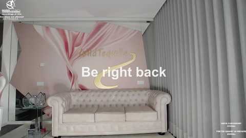 Media: Video of a stylish living room with a beige tufted couch, pink floral wallpaper, gray curtains, and a \"Be right back\" overlay.