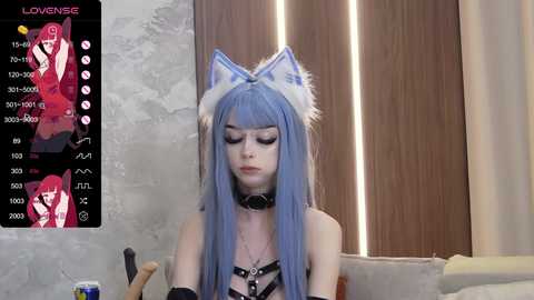 Media: Video of a pale, slender woman with long blue hair and cat ears, wearing a black leather harness and choker. She sits on a beige couch against a wooden backdrop.