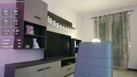 Media: Video of a modern living room with a flat-screen TV, white decorative figurines, a blue armchair, and white curtains with green leaf patterns.