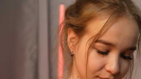 Media: A close-up video of a young woman with light brown hair, wearing a delicate necklace and a faint smile, set against a blurred, warm-toned background.